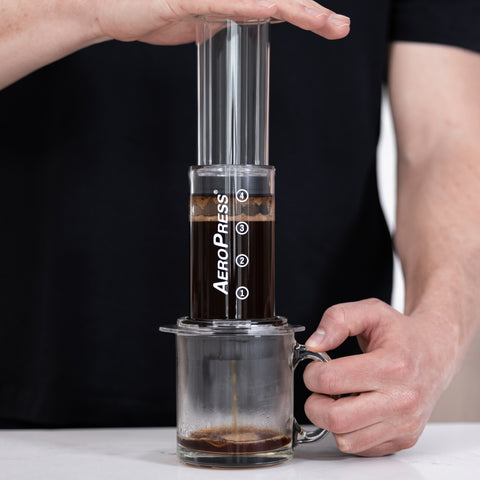 A man in a black t-shirt with one hand holding a clear glass mug and the other hand pushing down on a clear AeroPress.