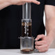 A man in a black t-shirt with one hand holding a clear glass mug and the other hand pushing down on a clear AeroPress.